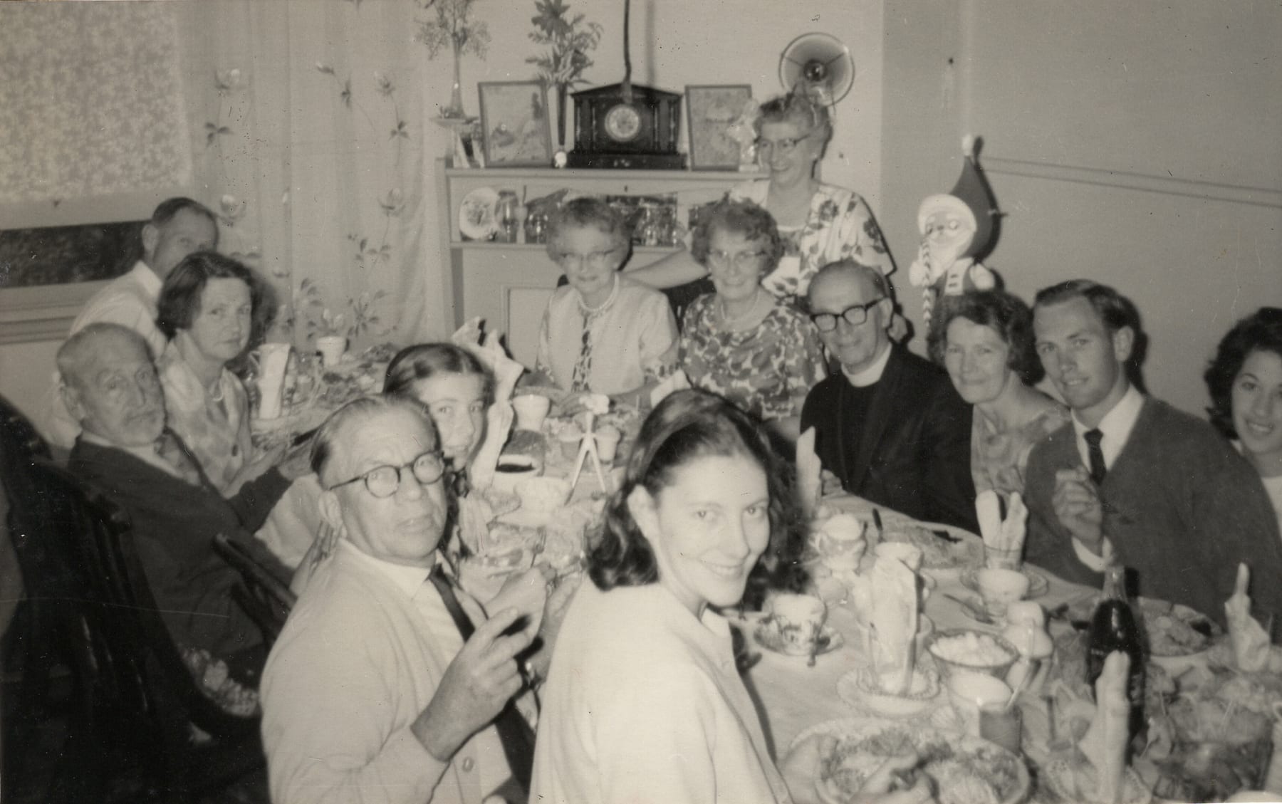 A dozen people crowded around a laden table indoors at Christmas, a Santa figure and a mantel clock on the sideboard behind; an older man in a clerical collar and dark-framed glasses sits among them at right.