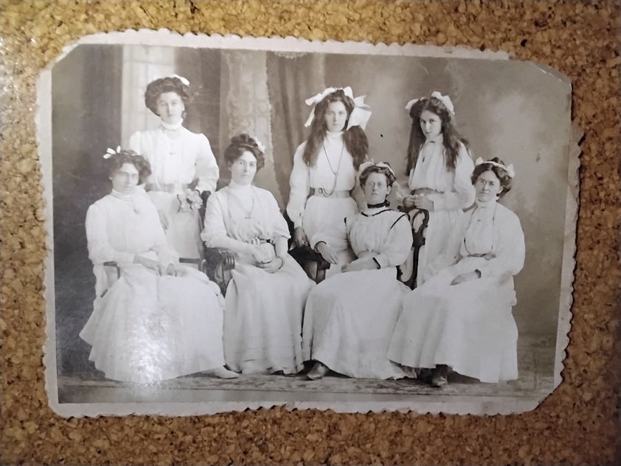 Seven young women in white high-necked dresses posed in a photographer’s studio — three standing behind, four seated in front, the two youngest wearing their hair loose with large white bows.