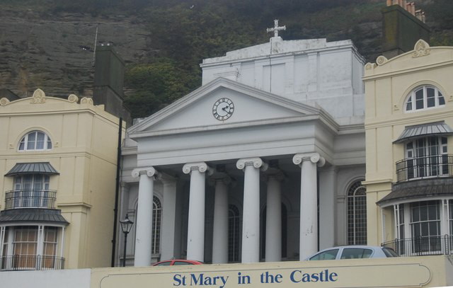 A white Greek-revival church front with a pillared portico and a clock set in the pediment, built into the cliff between two cream Regency terraces; a sign below reads “St Mary in the Castle”.