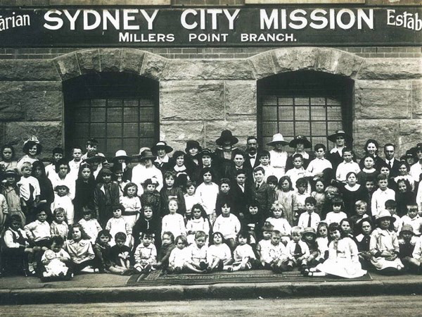 A large group of about eighty children and a dozen adults posed in rows on the steps and footpath outside a stone building; lettering above the windows reads “Sydney City Mission — Millers Point Branch”.