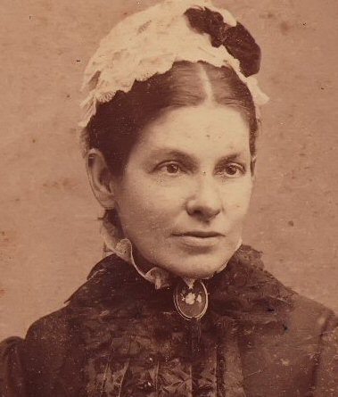 Sepia studio portrait of a woman in a dark high-necked dress with a brooch at the throat, wearing a pale lace cap.