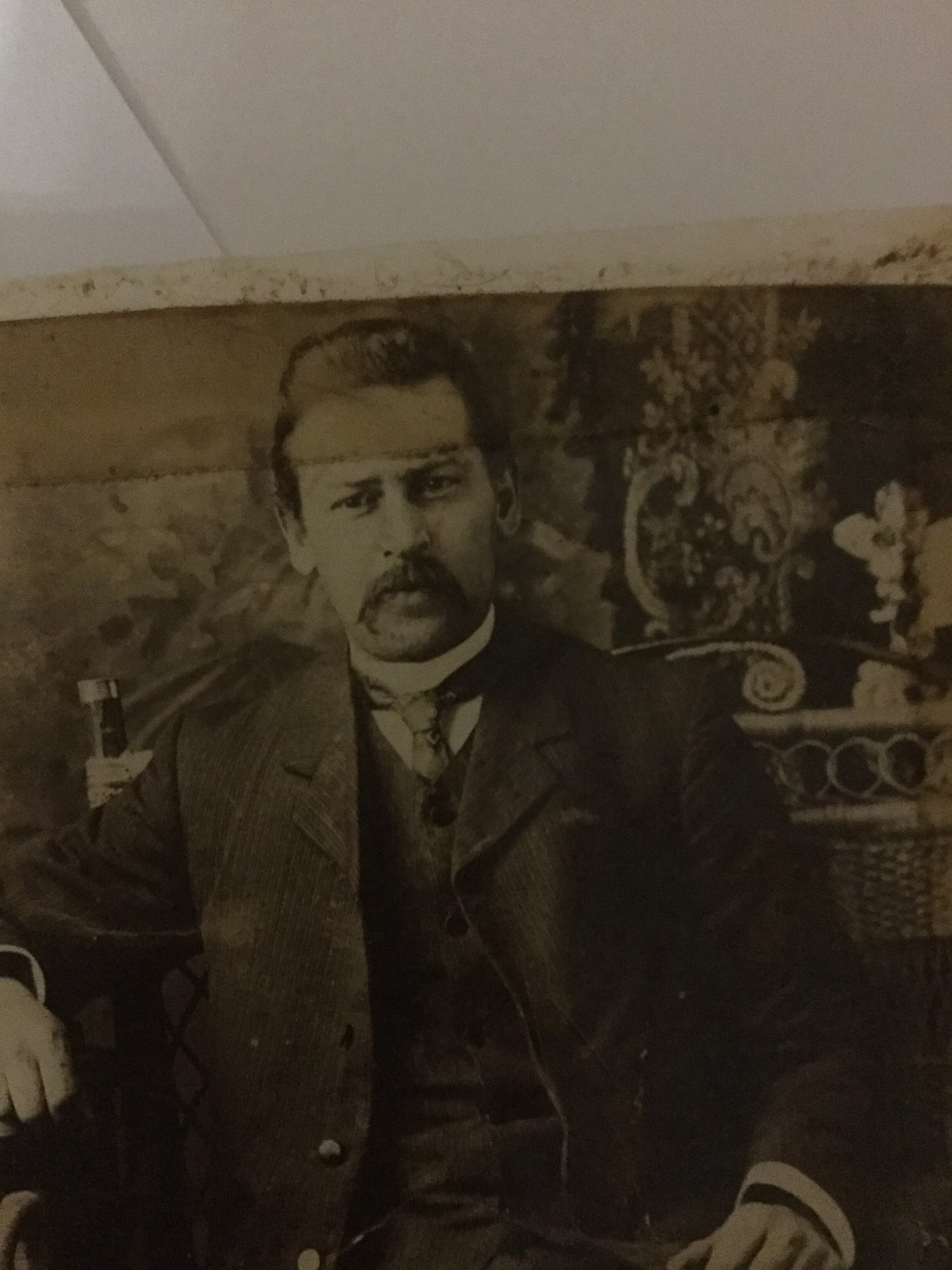 Studio half-length portrait of a moustached man in a sack suit and high collar, seated before a patterned backdrop.